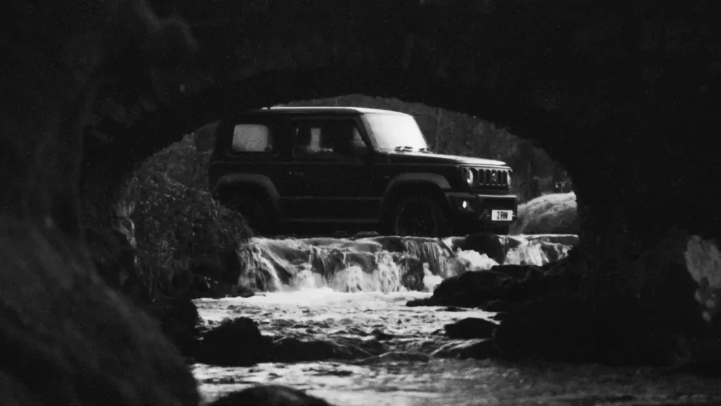 Suzuki Jimny under a stone bridge by a waterfall
